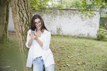 Junge Frau mit Smartphone lehnt an einem Baumstamm - FMKF03122