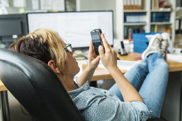 Frau liest Nachrichten auf dem Smartphone mit den Füßen auf dem Schreibtisch - TCF05165