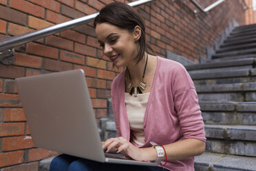Lächelnde Frau sitzt auf einer Treppe und arbeitet mit einem Laptop - BOYF00597