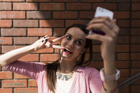 Porträt einer jungen Frau mit Siegeszeichen, die ein Selfie mit ihrem Smartphone macht, lizenzfreies Stockfoto