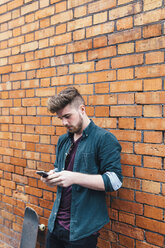 Young skateboarder leaning against brick wall looking at his cell phone - BOYF00602