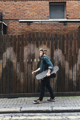 Young skateboarder walking on pavement listening music with headphones - BOYF00606