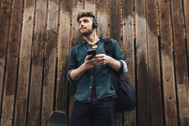 Junger Skateboarder vor einem Holzzaun, der mit Kopfhörern Musik hört - BOYF00608