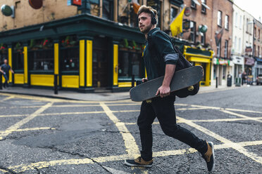 Irland, Dublin, junger Mann mit Kopfhörern und Skateboard beim Überqueren der Straße - BOYF00613