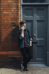 Junger Mann am Telefon, gegen die Eingangstür gelehnt - BOYF00614