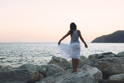 Rückenansicht einer Frau, die bei Sonnenuntergang auf einem Felsen vor dem Meer steht - GEMF01140