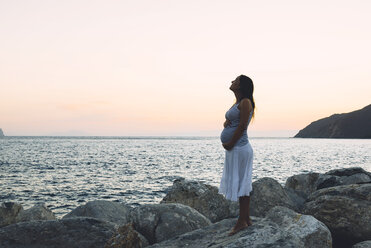 Pregnant woman standing on a rock in front of the sea by sunset - GEMF01141