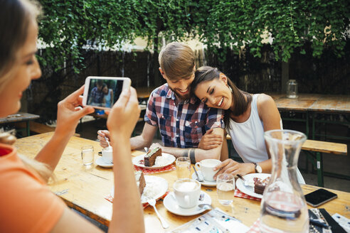 Woman taking cell phone picture of couple sitting outdoors with coffee and cake - AIF00382