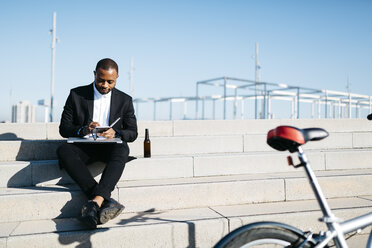 Businessman sitting on stairs with bottle of beer, notebook and laptop - JRFF00929