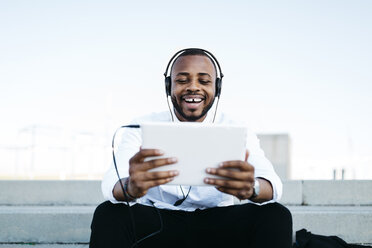 Happy man sitting on stairs wearing headphones looking at tablet - JRFF00939