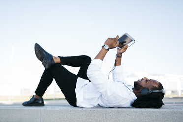 Smiling man lying on the floor wearing headphones looking at tablet - JRFF00941