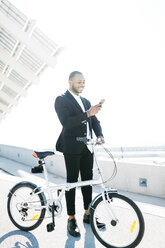 Smiling businessman with bicycle looking at cell phone - JRFF00948