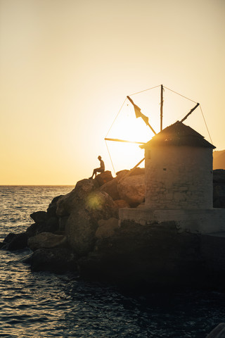 Griechenland, Amorgos, Aegialis, Silhouette eines Mannes, der in der Nähe einer Windmühle bei Sonnenuntergang sitzt, lizenzfreies Stockfoto