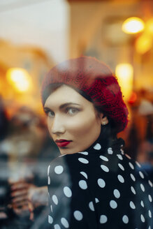 Portrait of young woman wearing red hat looking through window of a pub in the evening - LCUF00058