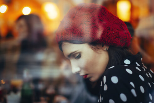 Porträt einer jungen Frau mit rotem Hut hinter der Fensterscheibe einer Kneipe am Abend - LCUF00059