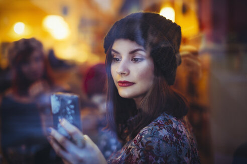 Portrait of young woman with smartphone behind windowpane of a pub in the evening - LCUF00061