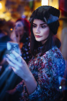 Portrait of young woman with smartphone behind windowpane of a pub in the evening - LCUF00063