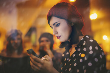 Junge Frau mit rotem Hut hinter der Fensterscheibe einer Kneipe am Abend - LCUF00065