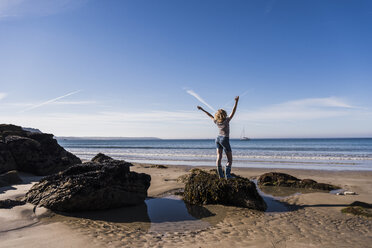 Frankreich, Halbinsel Crozon, junges Mädchen steht auf einem Felsen am Strand und hebt die Arme - UUF08642