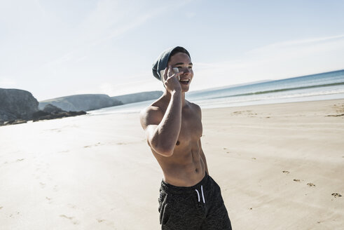 Frankreich, Halbinsel Crozon, glücklicher junger Mann mit Handy am Strand - UUF08666
