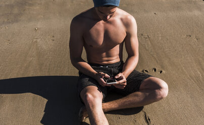 Junger Mann mit Mobiltelefon am Strand sitzend - UUF08667