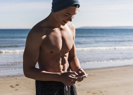 Frankreich, Halbinsel Crozon, lächelnder junger Mann mit Mobiltelefon am Strand - UUF08668