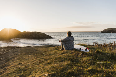 Frankreich, Bretagne, Halbinsel Crozon, Frau sitzt bei Sonnenuntergang mit Surfbrett an der Küste - UUF08682