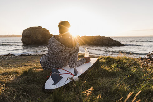 France, Bretagne, Crozon peninsula, woman sitting at the coast at sunset with surfboard - UUF08684