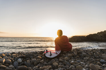 France, Bretagne, Crozon peninsula, woman sitting on stony beach at sunset with surfboard - UUF08688