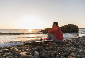 Frankreich, Bretagne, Halbinsel Crozon, Frau sitzt bei Sonnenuntergang mit Surfbrett am steinigen Strand - UUF08689