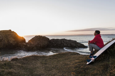 Frankreich, Bretagne, Halbinsel Crozon, Frau sitzt bei Sonnenuntergang mit Surfbrett an der Küste - UUF08695