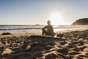 Frankreich, Bretagne, Halbinsel Crozon, Frau am Strand sitzend bei Sonnenuntergang mit Surfbrett - UUF08701