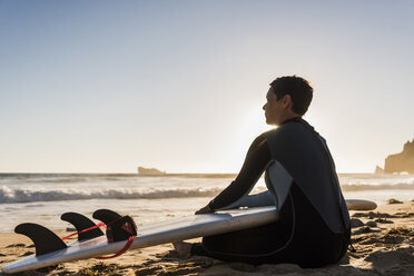 Frankreich, Bretagne, Halbinsel Crozon, Frau am Strand sitzend bei Sonnenuntergang mit Surfbrett - UUF08702