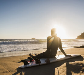 Frankreich, Bretagne, Halbinsel Crozon, Frau am Strand sitzend bei Sonnenuntergang mit Surfbrett - UUF08707