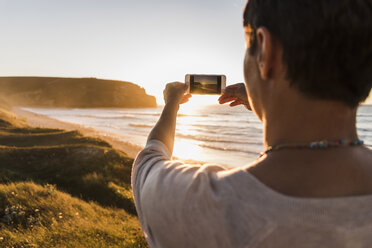 France, Bretagne, Crozon peninsula, woman at the coast at sunset taking picture with tablet - UUF08719