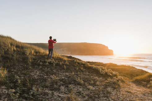 Frankreich, Bretagne, Halbinsel Crozon, Frau an der Küste bei Sonnenuntergang fotografiert mit Tablet - UUF08721