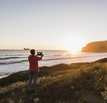 Frankreich, Bretagne, Halbinsel Crozon, Frau an der Küste bei Sonnenuntergang fotografiert mit Tablet - UUF08722
