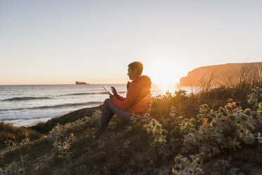 Frankreich, Bretagne, Halbinsel Crozon, Frau sitzt bei Sonnenuntergang an der Küste und benutzt Tablet - UUF08723