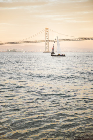 USA, San Francisco, Segelboot gegen Bay Bridge bei Sonnenuntergang, lizenzfreies Stockfoto