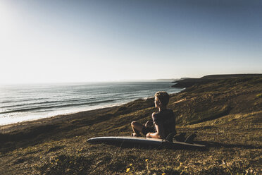 Jugendlicher mit Surfbrett am Strand sitzend und auf das Meer blickend - UUF08757