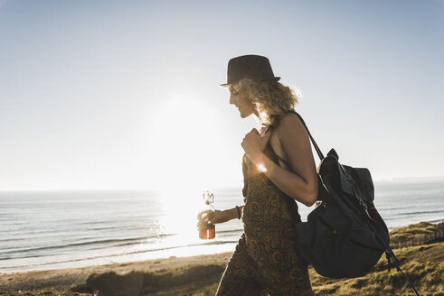 Blond teenage girl with backpack and beverage at seaside in the evening twilight - UUF08773