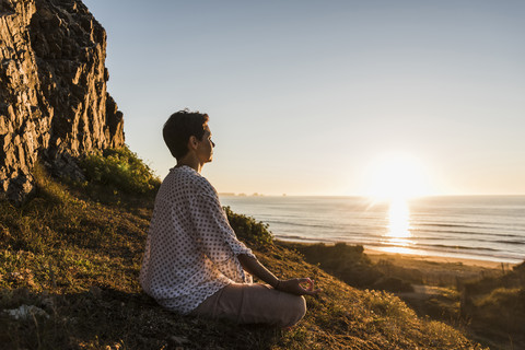 Frau meditiert auf einer Klippe bei Sonnenuntergang, lizenzfreies Stockfoto