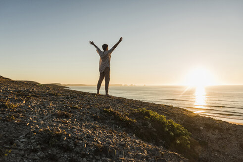 Frau mit erhobenen Armen auf einem Hügel stehend bei Sonnenuntergang mit Blick auf das Meer - UUF08804