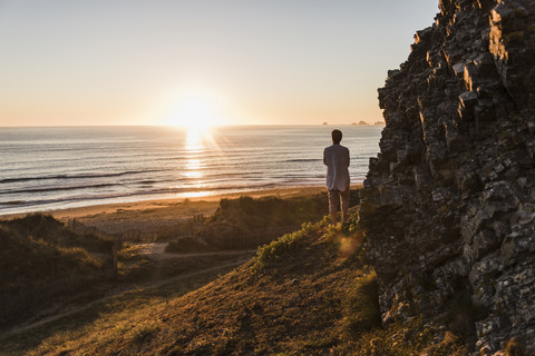 Rückenansicht einer Frau, die auf einer Klippe steht und den Sonnenuntergang beobachtet, lizenzfreies Stockfoto