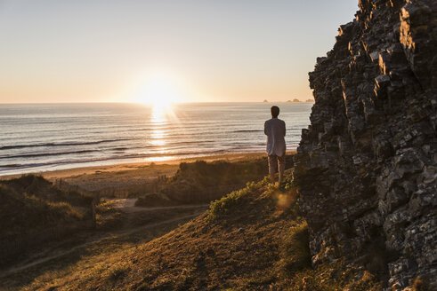 Rückenansicht einer Frau, die auf einer Klippe steht und den Sonnenuntergang beobachtet - UUF08806
