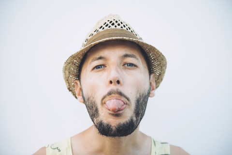 Porträt eines bärtigen Mannes mit Strohhut, der seine Zunge vor einem hellen Hintergrund herausstreckt, lizenzfreies Stockfoto