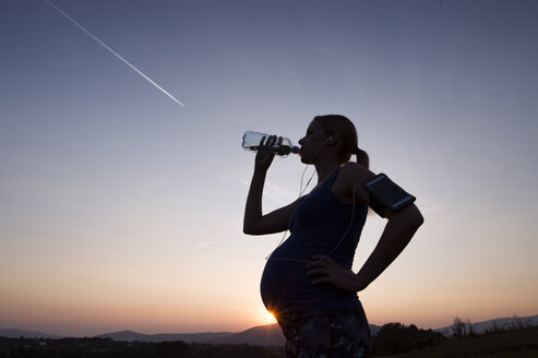 Silhouette einer schwangeren Frau, die bei Sonnenuntergang Wasser aus einer Flasche trinkt - HAPF01011