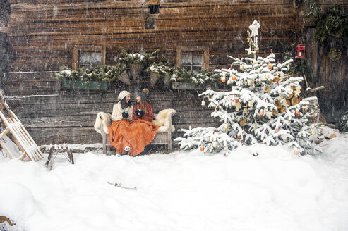 Friends sitting on bench by Christmas tree in front of mountain hut - HHF05432