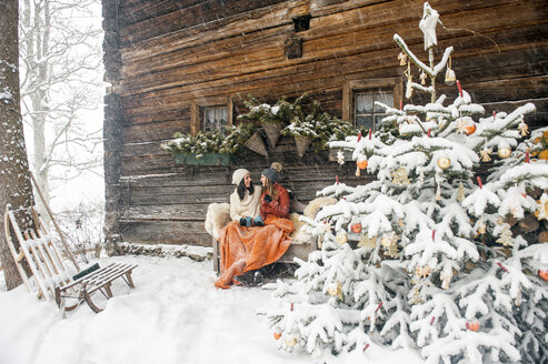 Friends sitting on bench by Christmas tree in front of mountain hut - HHF05433