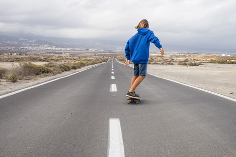 Spanien, Teneriffa, Rückansicht eines Jungen, der auf einer leeren Landstraße Skateboard fährt, lizenzfreies Stockfoto